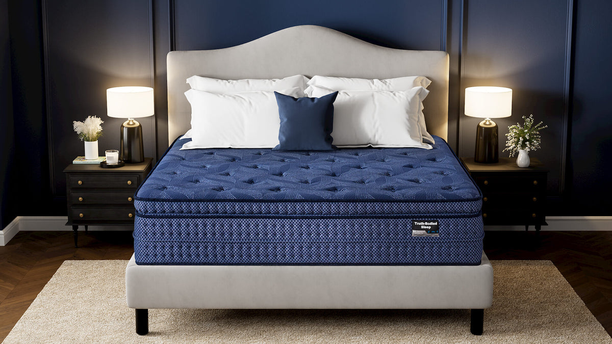 A luxury mattress shown on a bed in a styled bedroom. The mattress has a deep blue tufted top and a patterned blue side panel, set on a light gray upholstered bed frame with white and navy pillows against a dark navy paneled wall.