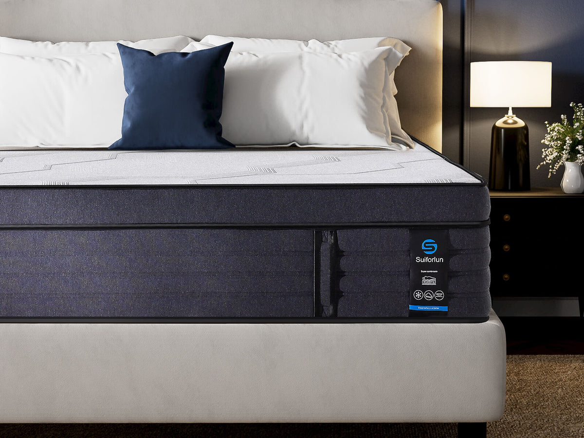 A Superfun mattress shown in a styled bedroom setting with white and navy pillows, a headboard, a bedside lamp, and a small vase of flowers.