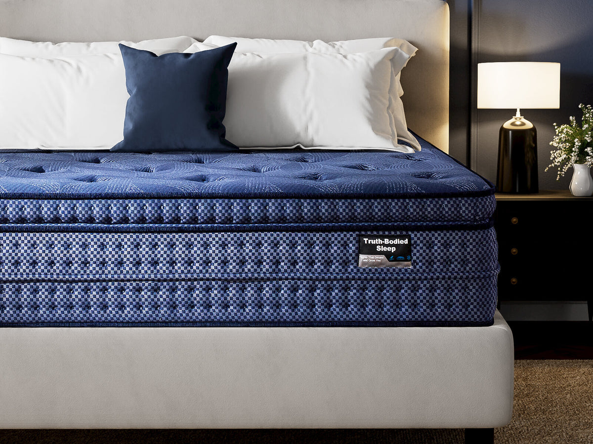 A luxury mattress shown in a styled bedroom setting, featuring a deep blue patterned mattress with a tufted top and a brand label on the side, set against a neutral gray headboard and bedside table with a lamp.