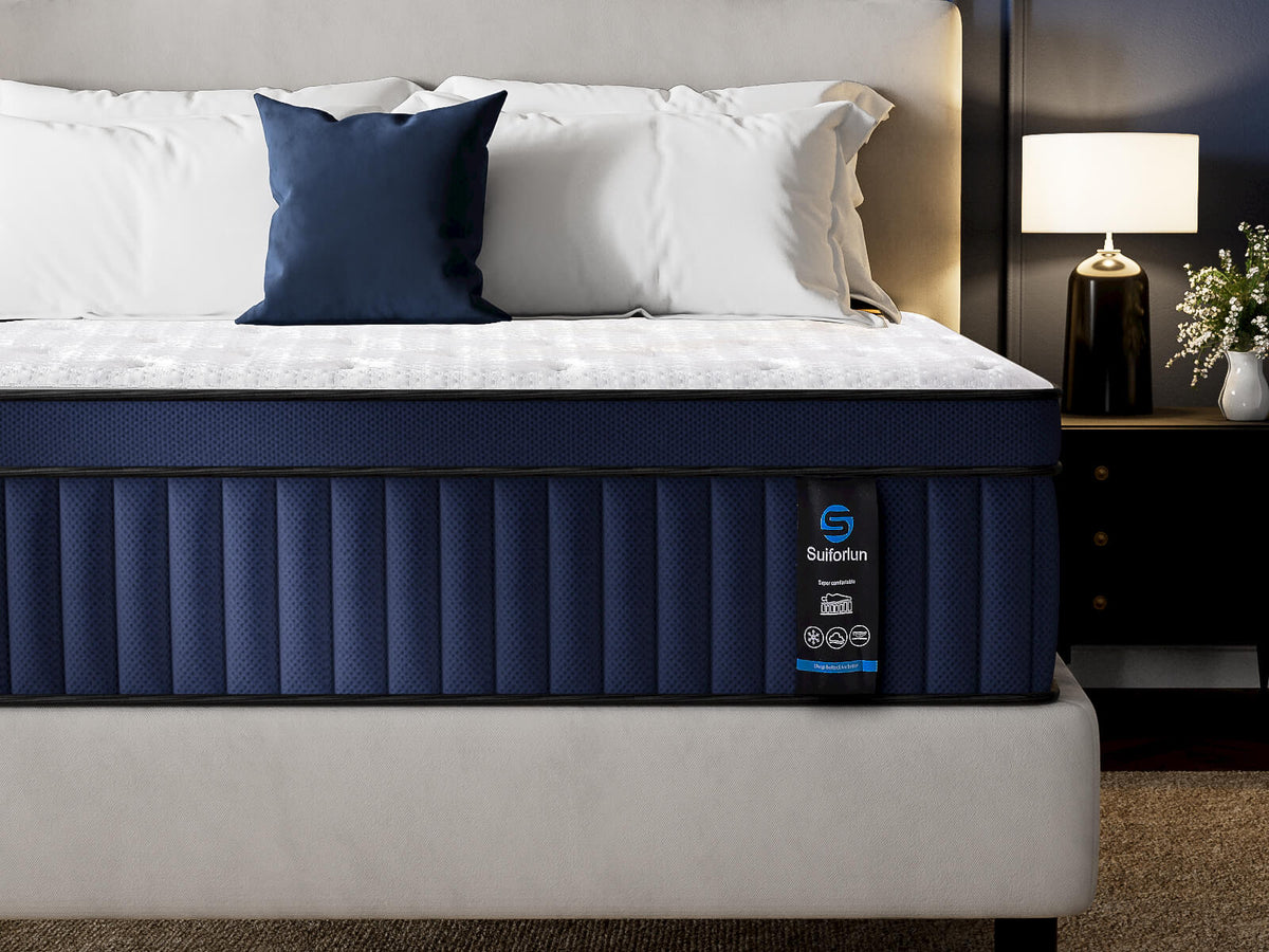 A bed dressed with white bedding and a navy blue accent pillow, featuring a dark navy upholstered mattress with a visible Suburban label. The bed is staged in a bedroom setting with a headboard, bedside table, lamp, and flowers.
